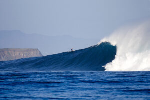 Surfer surfing a large big wave blue marine with a cliff, mountains and mystic sky in backgraund 