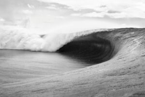 Black and white photo of a wave curling over itself with a cloudy sky.