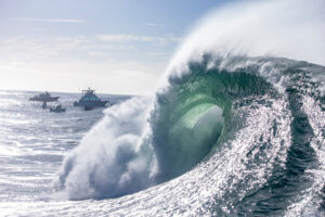 A powerful wave with a green curl forming in the center, with a boat and a small wave in the background.