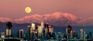 Luxury wall fine art of Downtown Los Angeles skyline at sunset with glowing skyscrapers, snow-capped mountains, and a rising full moon by Fred Pompermayer.